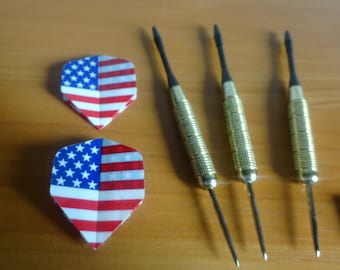 May include: A set of darts with American flag-themed flights. The flights are red, white, and blue, and the darts have gold-colored barrels and black tips. The darts are arranged on a wooden surface.