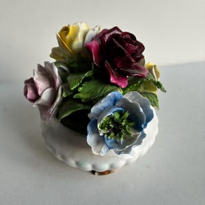 Pode incluir: Uma estatueta decorativa de porcelana apresentando um grupo de flores multicoloridas, incluindo uma rosa vermelha escura, uma rosa rosa pálido, uma rosa amarela e uma anémona azul, dispostas numa pequena taça de porcelana branca com borda festonada.