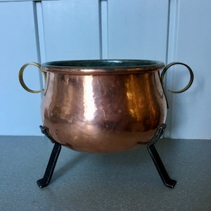 Könnte beinhalten: Ein Kupfertopf mit Messinggriffen und schwarzen Metallbeinen. Der Topf hat eine gehämmerte Oberfläche und steht auf einer grauen Oberfläche.