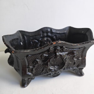 May include: A black cast iron planter, rectangular in shape, with decorative floral and scrollwork. It features a scalloped edge and stands on four small feet. The dark finish shows signs of wear and age.