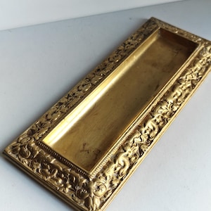 Może przedstawiać: Prostokątna, zabytkowa skrzynka na listy z mosiądzu z bogatymi detalami kwiatowymi i cherubinami. Skrzynka na listy ma długi, wąski otwór i ozdobną ramę. Mosiądz ma ciepły, złoty odcień.