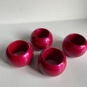 May include: Four round, fuchsia-colored napkin rings. Each ring has a smooth, glossy finish with subtle horizontal striations. The rings are evenly spaced against a white background, showcasing their vibrant color and design.