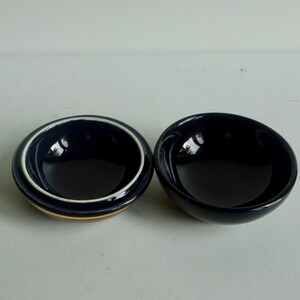 May include: Two small, round bowls. One bowl is dark blue with a white and gold rim. The other bowl is dark blue.