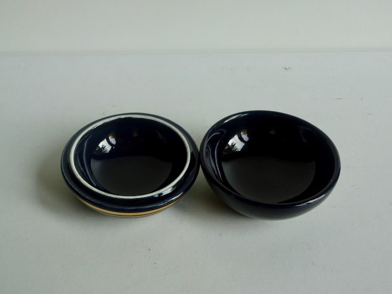 May include: Two small, round bowls. One bowl is dark blue with a white and gold rim. The other bowl is dark blue.