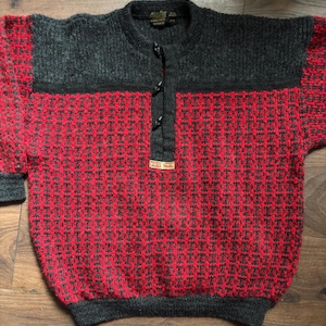 May include: A long-sleeve pullover sweater featuring a charcoal gray upper section and a vibrant red and charcoal gray patterned lower body. The sweater has a button-up placket with three black toggles and a small rectangular brand label at the base.