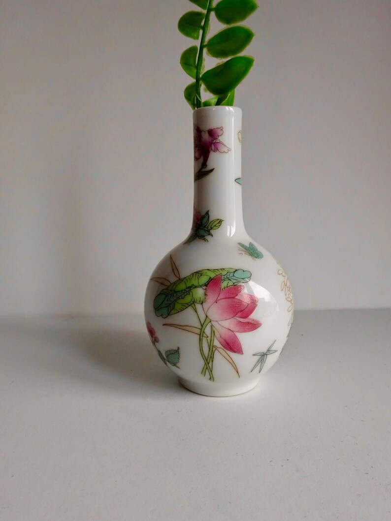 May include: A small white porcelain vase with a floral design featuring pink and green flowers. The vase has a round body and a narrow neck. A green leafy stem is in the vase.