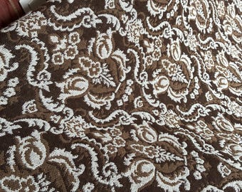 May include: A woven textile featuring a dense, repeating floral and scrollwork pattern in contrasting shades of dark brown and off-white. The fabric has a textured appearance with a visible weave and a decorative fringe along the bottom edge.
