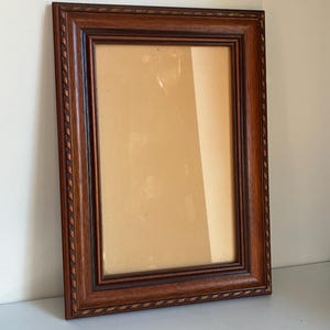 Op de afbeelding: Een rechthoekige houten fotolijst met een donkerbruine afwerking. Het frame heeft een decoratief touwachtig patroon langs de randen en een lichtere bruine binnenrand. Het frame is leeg, met een tan achterkant zichtbaar.