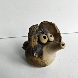 Op de afbeelding: Een klein, rond, met de hand gevormd keramieken beeldje met een textuurrijke, gevlekte bruine en beige afwerking. Het object heeft twee opvallende, uitstekende ronde ogen en een lange, taps toelopende snuit met twee kleine gaatjes aan het uiteinde.