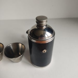 Peut inclure: Un shaker à cocktail vintage avec un corps noir et un dessus et un couvercle argentés brillants. Deux petites tasses à mesurer en métal argenté sont visibles à gauche du shaker. Le shaker a un design classique, adapté au mélange de boissons.