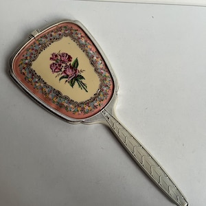Puede incluir: Un espejo de mano plateado con un mango alargado que presenta un patrón de hojas. La parte posterior del espejo muestra un panel central de color crema con un ramo de rosas rosas, rodeado por un borde decorativo de pequeñas flores coloridas.