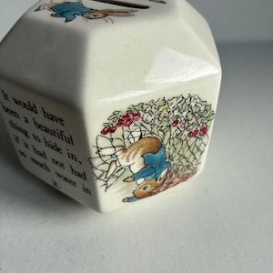 May include: Cream-colored ceramic money bank with a hexagonal shape. The bank features illustrations of a rabbit in a blue jacket and text from a children's story. A slot is visible on the top for inserting money.