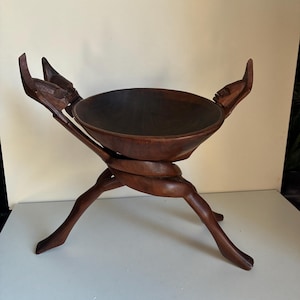 Peut inclure: Un bol en bois sculpté à la main, de couleur marron foncé, avec un bassin rond et profond. Le bol est soutenu par un trépied avec des jambes entrelacées et deux figures sculptées sur les côtés.