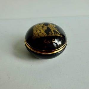 May include: A small, round, black ceramic trinket box with a gold trim and a gold design of two figures on the lid. The lid is inscribed with the word "Corfu".