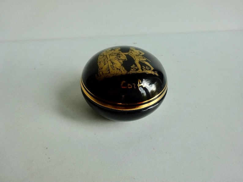 May include: A small, round, black ceramic trinket box with a gold trim and a gold design of two figures on the lid. The lid is inscribed with the word "Corfu".