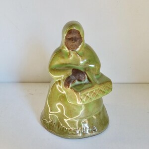 Può includere: Figurina in ceramica verde di una donna che indossa un mantello con cappuccio e tiene un cestino. La figurina è smaltata e ha un aspetto rustico. La base della figurina è incisa con la parola "Made in USA."