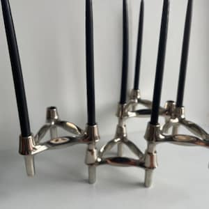 May include: A silver-toned metal candelabra with six black taper candles. The candelabra has a modern, geometric design with a series of interconnected arms and cylindrical candle holders. The candles are unlit and have white wicks.