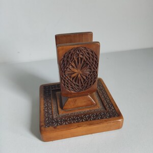 Könnte beinhalten: Ein Serviettenhalter aus Holz mit geometrischem Design. Der Halter hat eine quadratische Basis mit geschnitztem Rand, einen zentralen Sockel und zwei aufrechte Paneele mit einem geschnitzten, kugelförmigen Muster. Das Holz hat einen warmen, natürlichen Farbton.