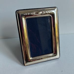 May include: A rectangular picture frame with a polished silver-tone metal border featuring an ornate embossed design at the top centre. The frame has a dark blue textured insert and a black velvet backing visible along the outer edges.
