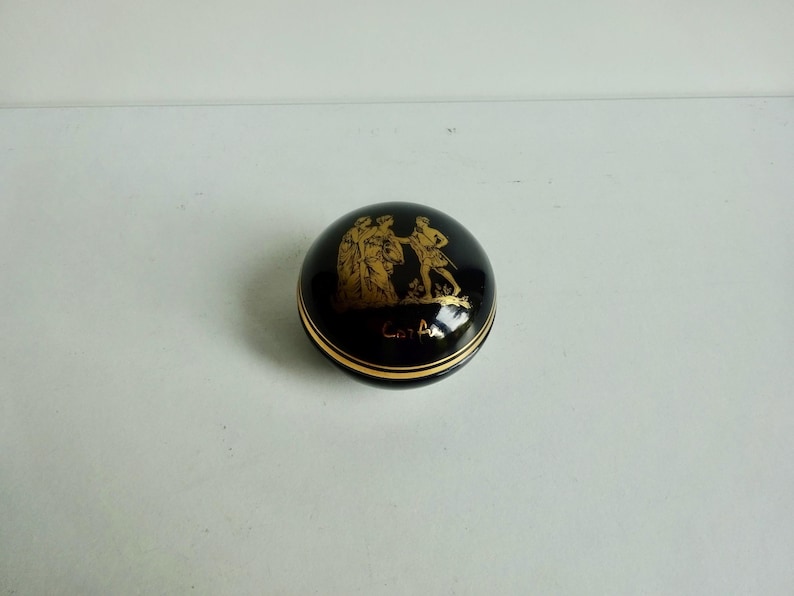 May include: A black and gold round trinket box with a gold image of three figures on the lid. The box is inscribed with the text "Art A".