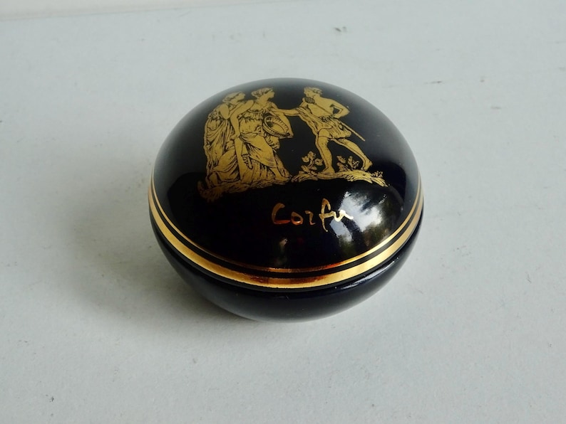 May include: A small, round, black ceramic trinket box with a gold trim and a gold image of three figures on the lid. The box is inscribed with the word "Corfu" in gold lettering.