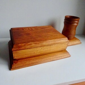 Op de afbeelding: Een houten doos met een scharnierend deksel en een aparte houten standaard. De doos is gemaakt van lichtbruin hout met een natuurlijke afwerking. De standaard is ook gemaakt van lichtbruin hout en heeft een eenvoudig ontwerp.