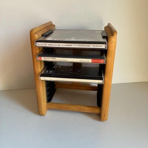 May include: A wooden CD rack with three shelves, each holding a CD case. The rack is made of light-coloured wood and has a simple, functional design. The CD cases are black and have white labels.