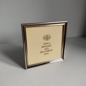 Könnte beinhalten: Ein silberfarbener, quadratischer Fotorahmen mit einem Perlenrand. Der Rahmen enthält eine cremefarbene Rückseite mit dem Text "Hallfield" und "To finest photography add a Spicer Hallfield frame."
