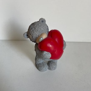Op de afbeelding: Een grijze teddybeerfiguur met een glanzend rood hart. De beer heeft een klein, rond, lichtgekleurd voorwerp om zijn nek. Het beeldje staat op een wit oppervlak.