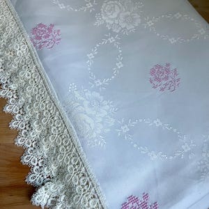 May include: A folded white tablecloth with a floral pattern and cream lace trim. The floral design features white roses and other flowers, with pink accents. The lace trim is along the edge of the tablecloth.