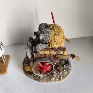 May include: A detailed figurine of two teddy bears in a hay cart. The cart has a red wheel and is filled with straw. The bears are grey and brown, and the scene is set on a base with small stones.