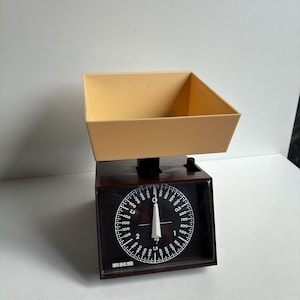 May include: A brown mechanical kitchen scale with a beige square plastic tray on top. The circular black dial features white markings, numbers, and the brand name EKS. A white needle points to the zero position on the scale face.