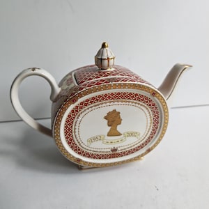 Op de afbeelding: Ovale keramische theepot met een rood en goud patroon. De theepot heeft een wit ovaal met een gouden silhouet van een koningin en de jaren 1952 en 2002. De handgreep en tuit zijn wit met gouden accenten. Een gouden knop bekroont het deksel.