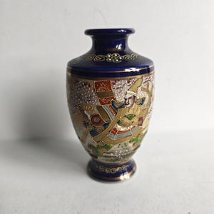 May include: A small ceramic vase with a deep blue neck and base featuring gold trim. The body displays a detailed hand-painted scene of a figure in traditional armor, accented with gold, red, and green colors against a textured, speckled background.