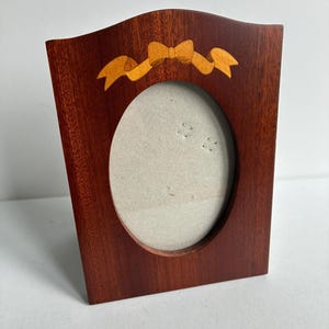 Puede incluir: Un marco de fotos de madera de color marrón rojizo oscuro con una abertura central ovalada. La parte superior del marco presenta un diseño decorativo de lazo de cinta incrustado en madera clara. El marco tiene un borde superior curvo.