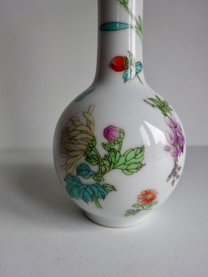 May include: A white porcelain vase with a narrow neck and a round body. The vase is decorated with a floral pattern in shades of pink, purple, green, yellow, and red.