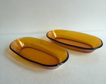 Op de afbeelding: Twee ovaalvormige schalen gemaakt van doorschijnend amberkleurig glas. Elke schaal heeft een gladde, gebogen rand en een ondiepe kom, met een consistente warme, goudbruine tint door het gehele glasmateriaal.