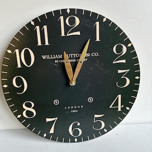 May include: A round wall clock with a dark green face, featuring large cream-colored serif numerals from 1 to 12. The center displays the text WILLIAM UTTON AND CO. 85 CHEAPSIDE ST. LONDON 1894. It has two gold-toned metal hands and a minute track.