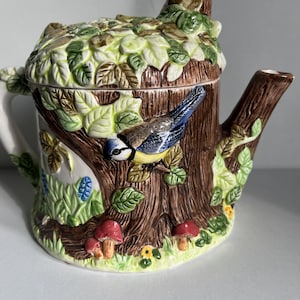 Op de afbeelding: Een keramische theepot met een grillig bosontwerp. De theepot heeft de vorm van een boomstam, met een blauwe en gele vogel die aan de zijkant zit. Het deksel is bedekt met groene bladeren en de basis heeft rode paddenstoelen en gele bloemen.