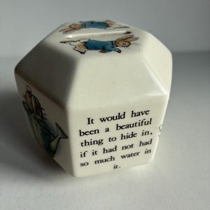 May include: Cream-colored ceramic money box with a multi-faceted design. The top features illustrations of a rabbit wearing a blue jacket. The side of the box has text that reads, "It would have been a beautiful thing to hide in, if it had not had so much water in it."