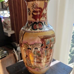 Könnte beinhalten: Eine hohe, verzierte Vase mit breitem Boden und ausgestelltem Rand. Die Vase zeigt detaillierte Kunstwerke, die Figuren in traditioneller Kleidung darstellen, mit goldenen Akzenten und einer warmen Farbpalette aus Rot-, Gelb- und Rosatönen. Die Vase ist wahrscheinlich aus Keramik.