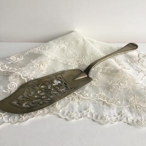 Puede incluir: Un servidor de pastel de plata con un diseño ornamentado y calado. El servidor está sobre un encaje blanco.
