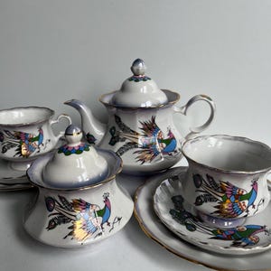 Op de afbeelding: Een porseleinen theeservies bestaande uit een theepot, een suikerpot met deksel en twee theekopjes met bijpassende schoteltjes. Elk stuk is wit met een parelmoer afwerking, goudkleurige randen en een kleurrijke pauwenillustratie in blauw, groen en roze.