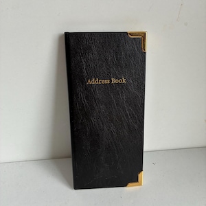 Op de afbeelding: Zwart adresboek met gouden hoekaccenten. De omslag heeft een getextureerde uitstraling en de woorden "Address Book" zijn in goud gedrukt. Het boek is rechthoekig en staat rechtop tegen een witte achtergrond.
