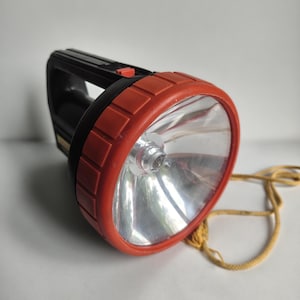 Könnte beinhalten: Eine schwarze und rote Taschenlampe mit einer großen, reflektierenden Linse. Die Taschenlampe hat einen schwarzen Griff und eine rote Lünette um die Linse. Ein gelbes Seil ist befestigt.