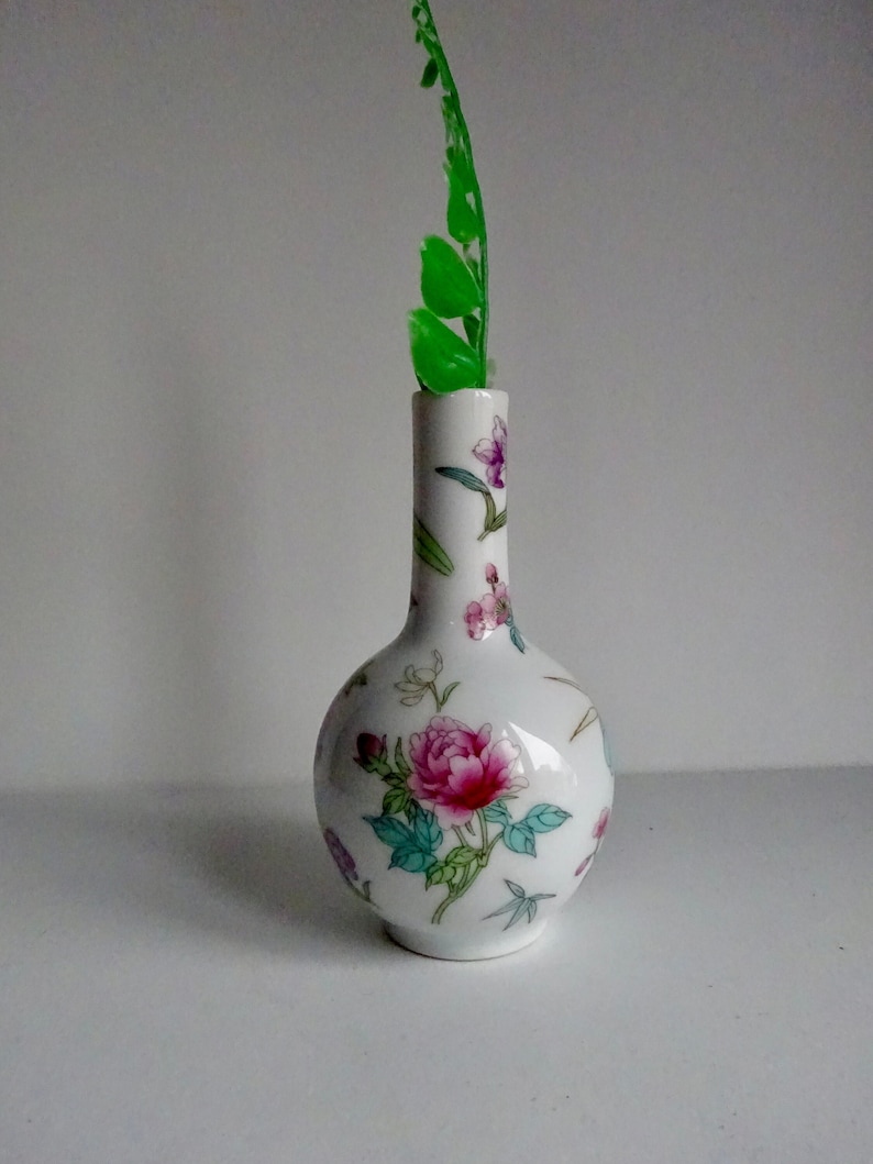 May include: A small white porcelain vase with a floral pattern in shades of pink, green, and blue. The vase has a round body and a narrow neck. A green leaf is placed in the vase.