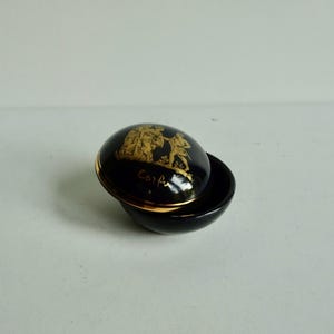 May include: A small, round, black ceramic trinket box with a gold rim and a gold design on the lid. The design features two figures in a scene with the word "Corfu" written below.