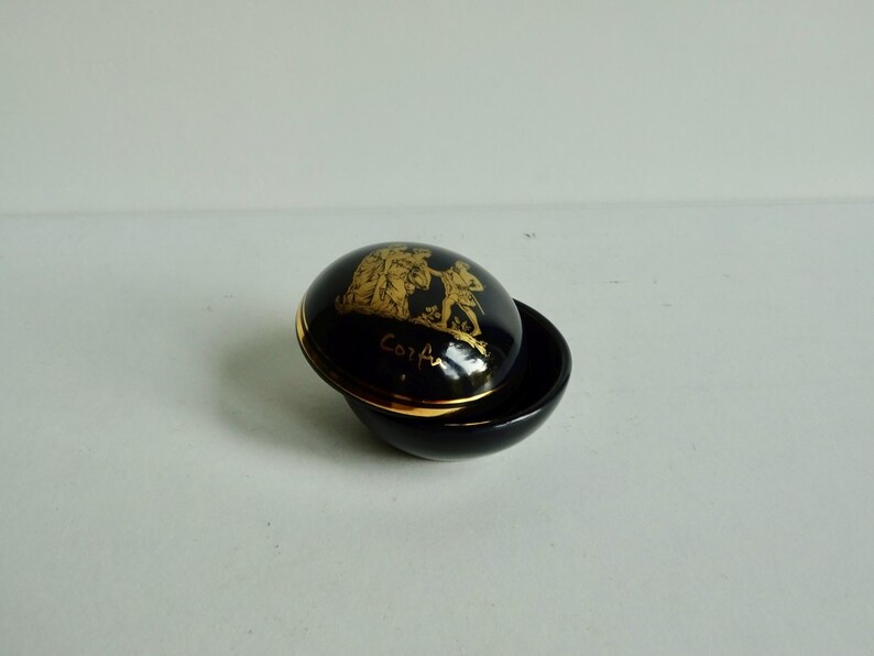 May include: A small, round, black ceramic trinket box with a gold rim and a gold design on the lid. The design features two figures in a scene with the word "Corfu" written below.