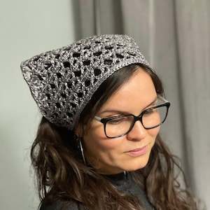 May include: A silver crochet headband with a delicate openwork pattern. The headband is worn by a person with long brown hair and glasses.