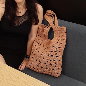 May include: A tan crochet tote bag featuring a repeating open-work geometric pattern with rectangular cutouts and two integrated shoulder straps. The bag is constructed from a textured, fibrous material and includes a small brown leather-like tag.